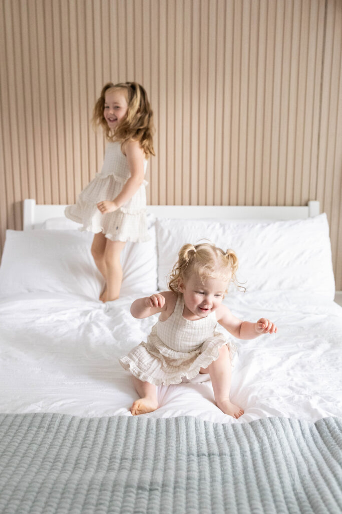 Big sisters jumping on the bed with newborn baby Taylor during an in-home lifestyle newborn photography session in Northern Virginia.